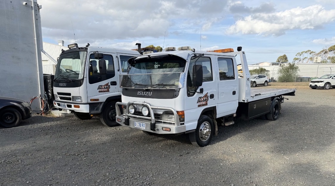 Two white Axis Towing Isuzu tilt-tray trucks parked in the yard